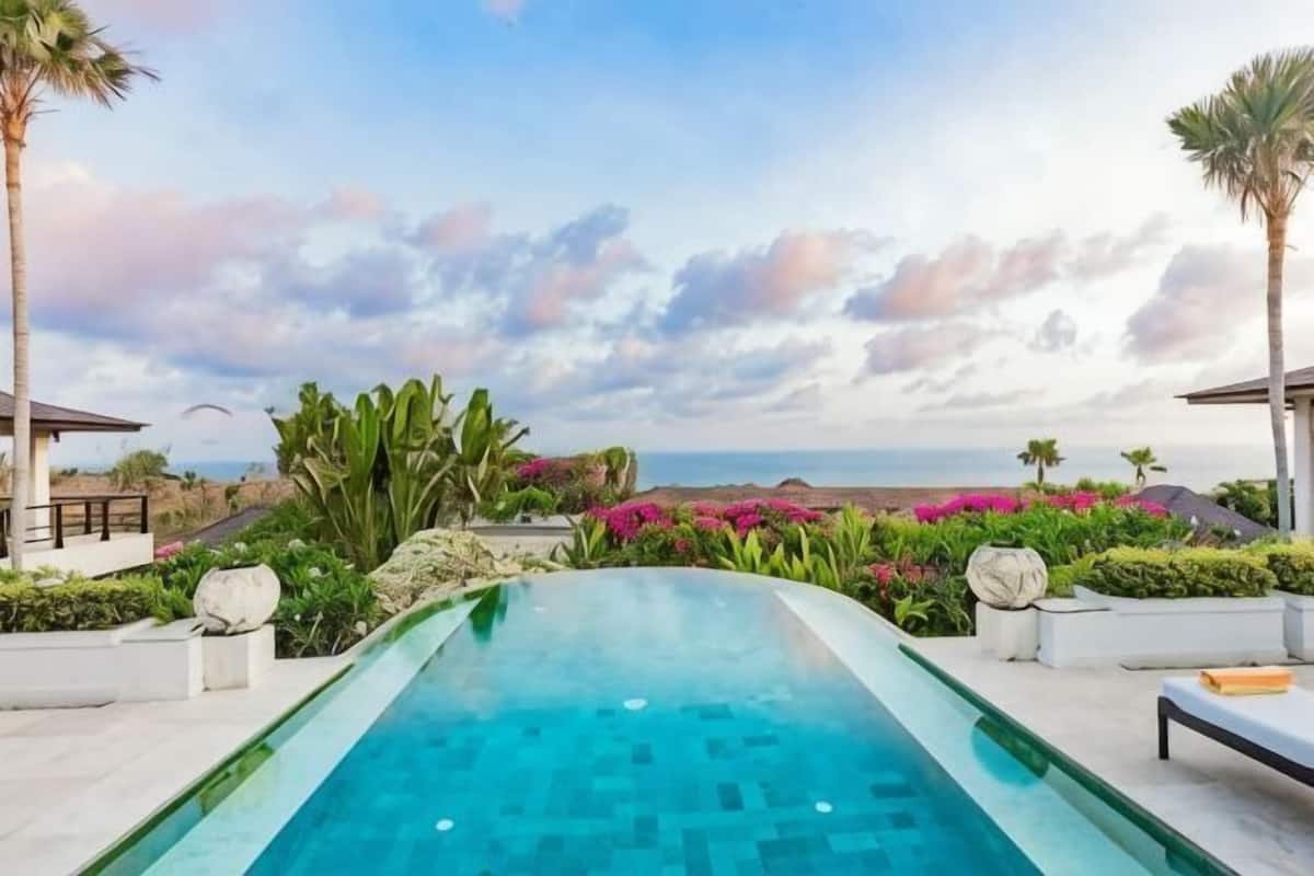 A swimming pool with a view of the ocean, surrounded by tropical plants and palm trees.