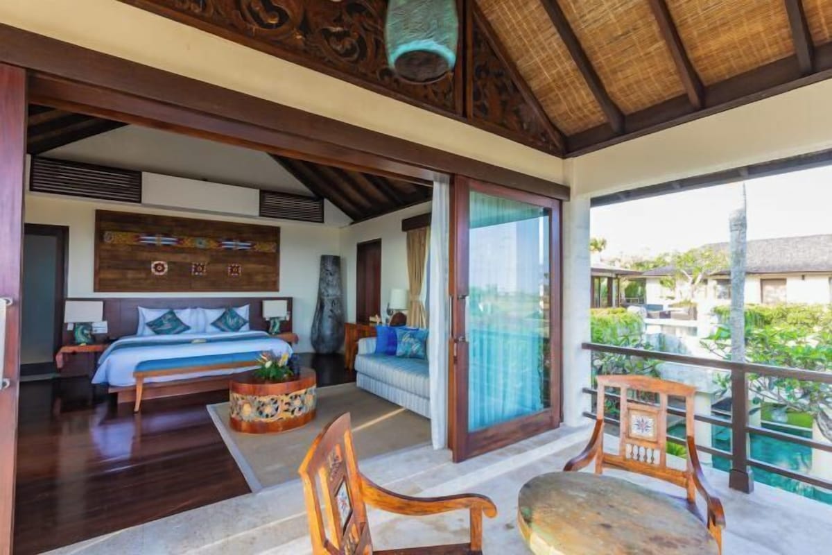 A spacious bedroom with a large bed, a wooden ceiling, and a balcony with a view of the pool and greenery.