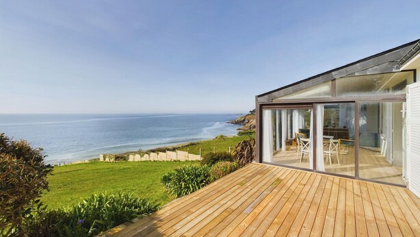Una casa moderna con una terraza de madera y un ventanal de vidrio que da a un área de comedor exterior con vista al océano.