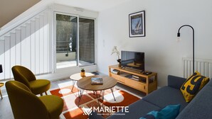 Ein modernes Wohnzimmer mit Sofa, Couchtisch und Fernseher auf einem Holzboden. Ein großes Fenster mit Jalousien, eine Treppe und eine hängende Leuchte sind ebenfalls zu sehen.