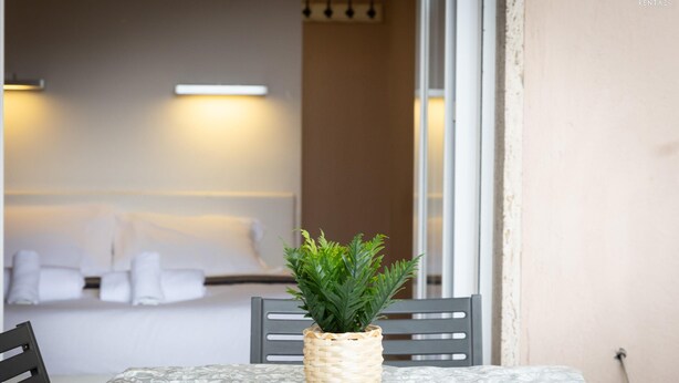 A dining table with a woven plant pot, two chairs, and a view of a room with a bed and towels.