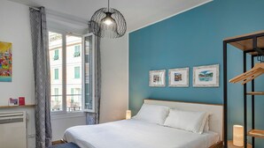 A bedroom with a bed, a bench, a window with curtains, a wall-mounted light fixture, and three framed pictures. - Art near Lomellini Street by Wonderful Italy (Genoa)