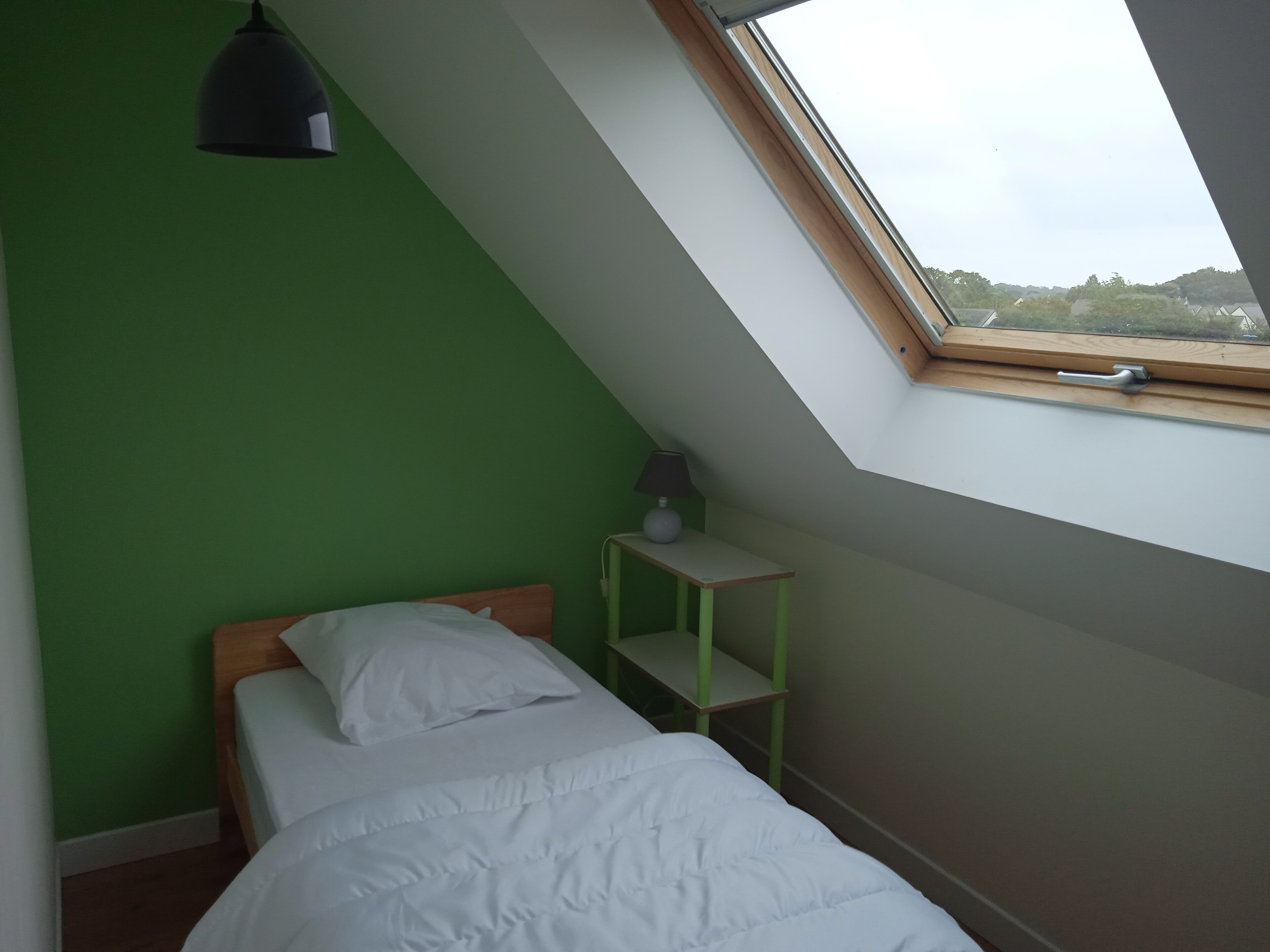 A room with a sloped ceiling, a bed with white bedding, a small table with a lamp, and a window with a view of greenery.