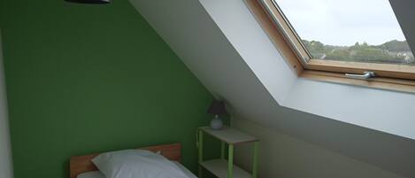 A room with a sloped ceiling, a bed with white bedding, a small table with a lamp, and a window with a view of greenery.