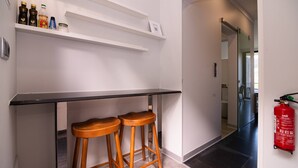 Une cuisine avec un plan de travail noir, deux tabourets en bois et une étagère blanche sur laquelle sont disposés divers objets.