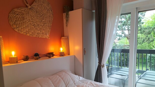 Un dormitorio con cama, estante con adornos, un colgador con forma de corazón y balcón con barandilla.