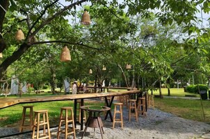 Un área de comedor al aire libre con mesas y taburetes de madera, rodeada de árboles y faroles colgantes.