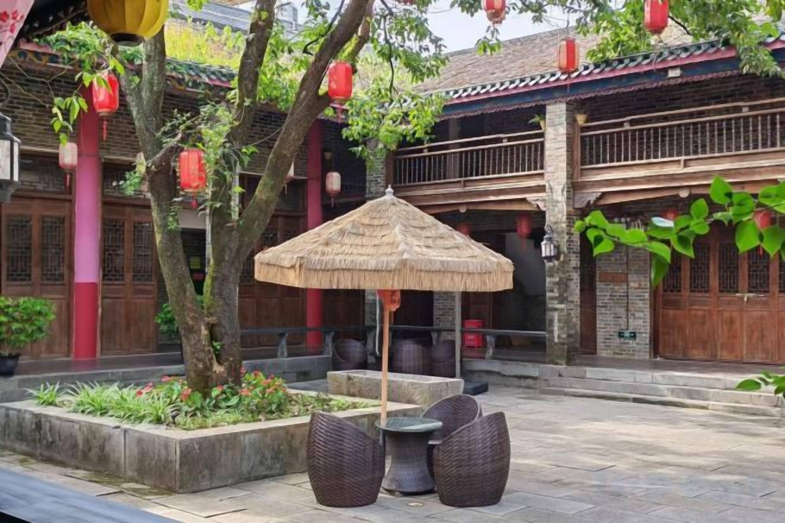 Un patio exterior con arquitectura tradicional, que incluye una sombrilla de paja, muebles de mimbre y faroles rojos.