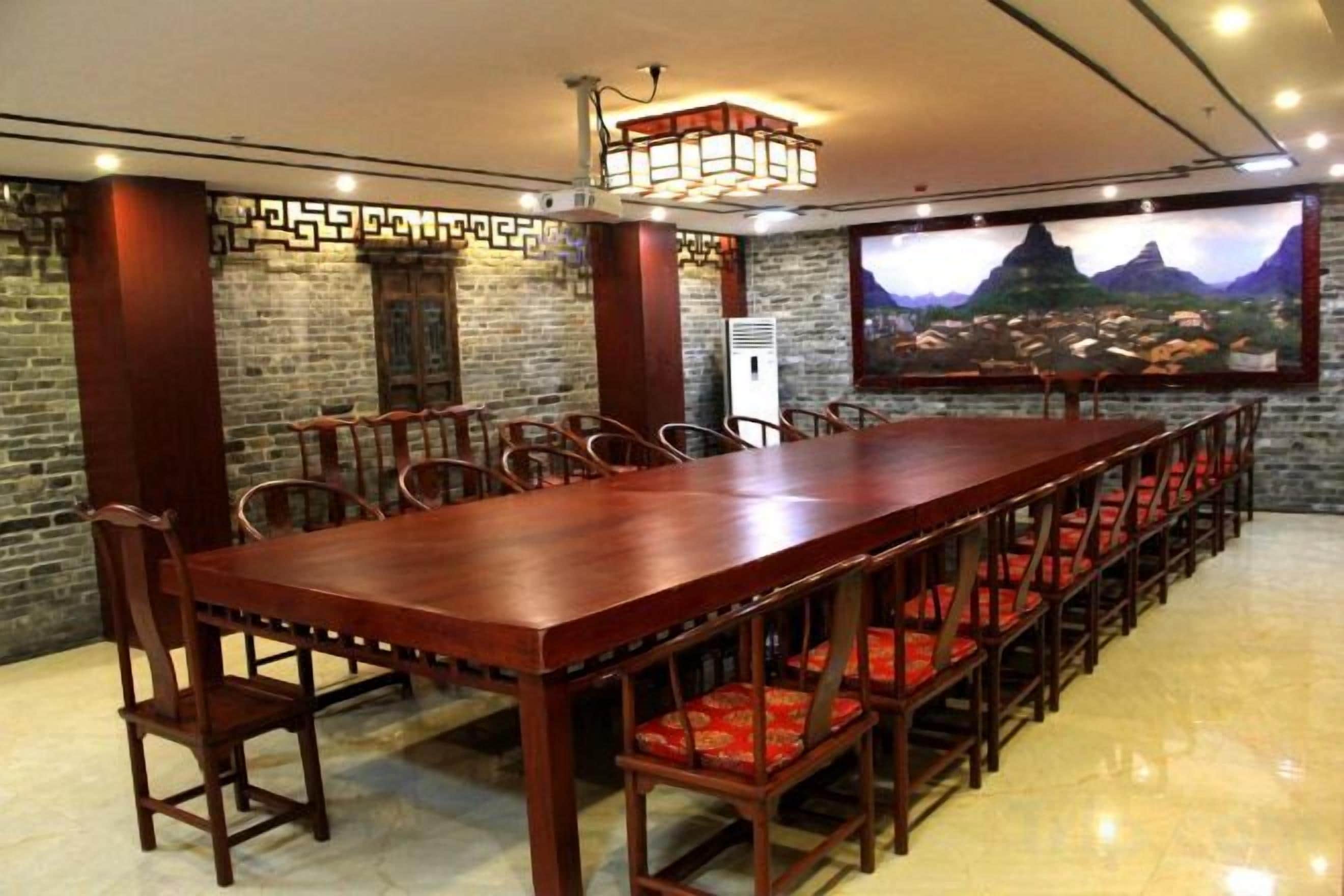 A long wooden dining table with red cushioned chairs, a chandelier, and a large wall-mounted TV displaying a scenic landscape.