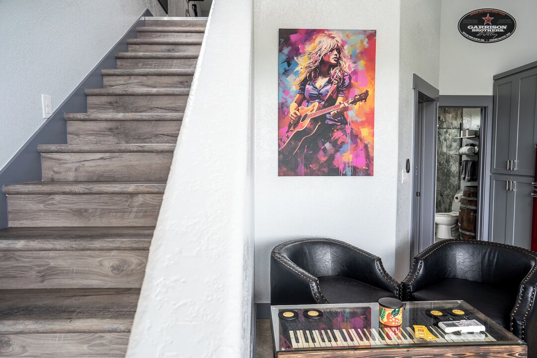 Een trap met houten treden, een zwarte leren bank, een glazen salontafel met een toetsenbord en platen, en een kleurrijk gitaar schilderij aan de muur.