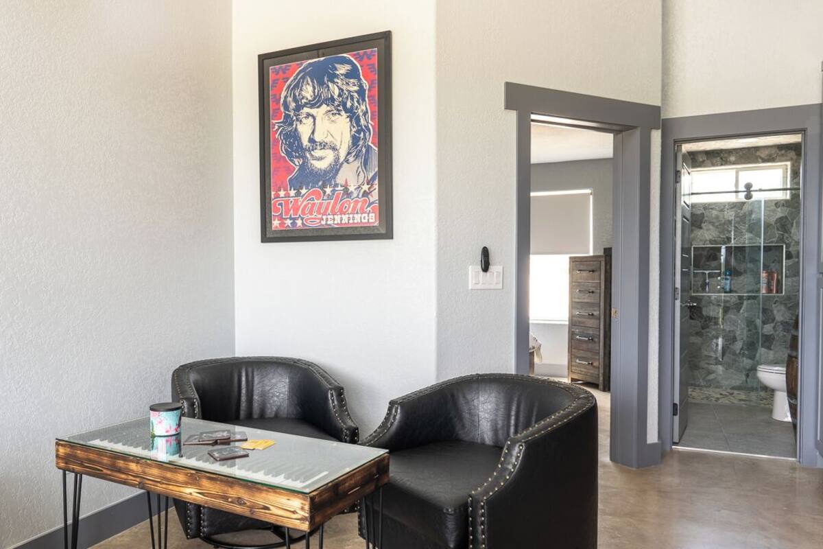 Ein Zimmer mit einem Glastisch, zwei Ledersesseln, einem gerahmten Poster an der Wand und einer Tür zu einem Badezimmer mit einer Steindusche.