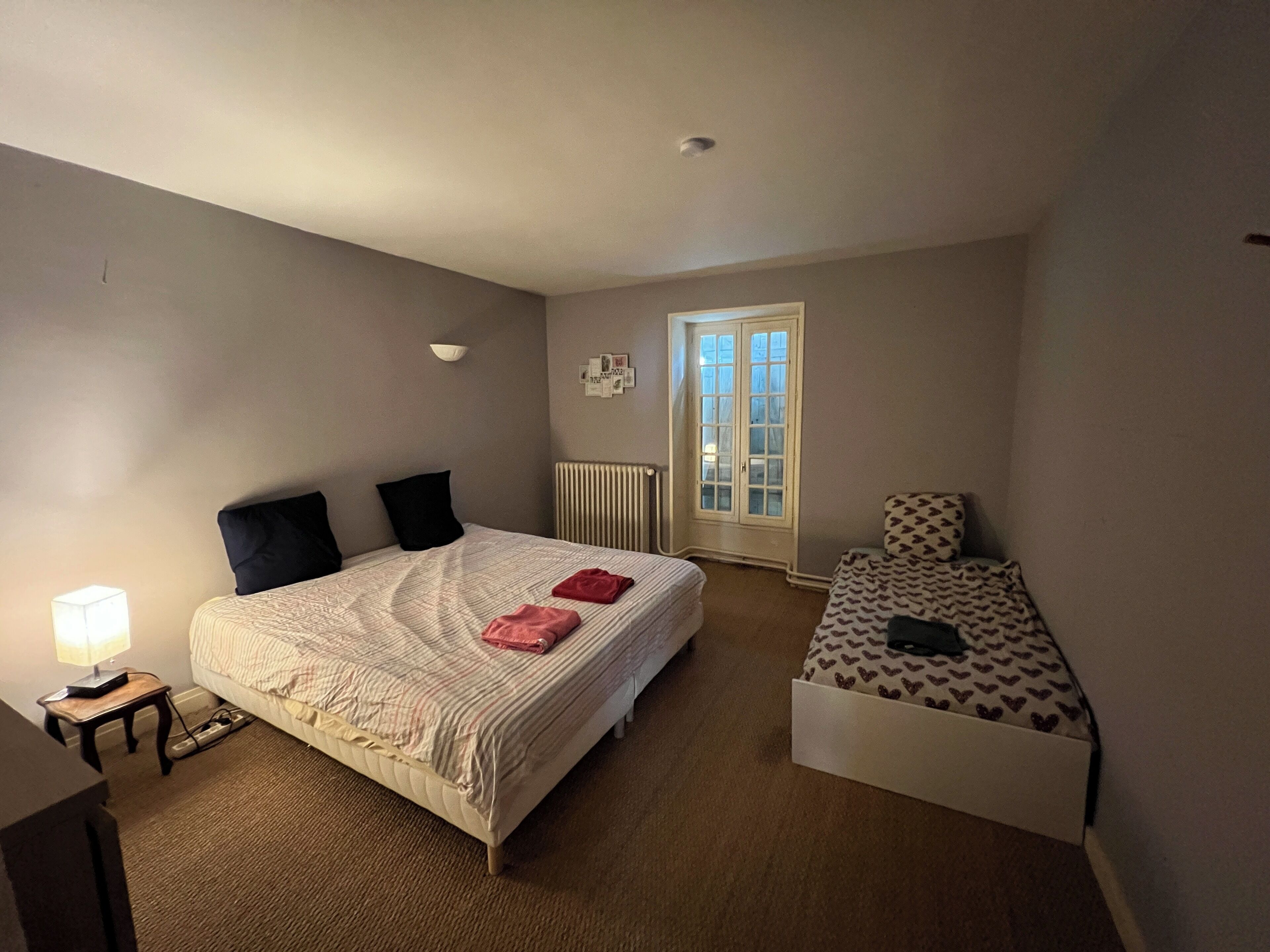 Una camera da letto con un letto grande, un letto più piccolo, un termosifone, una finestra e un comodino con una lampada.