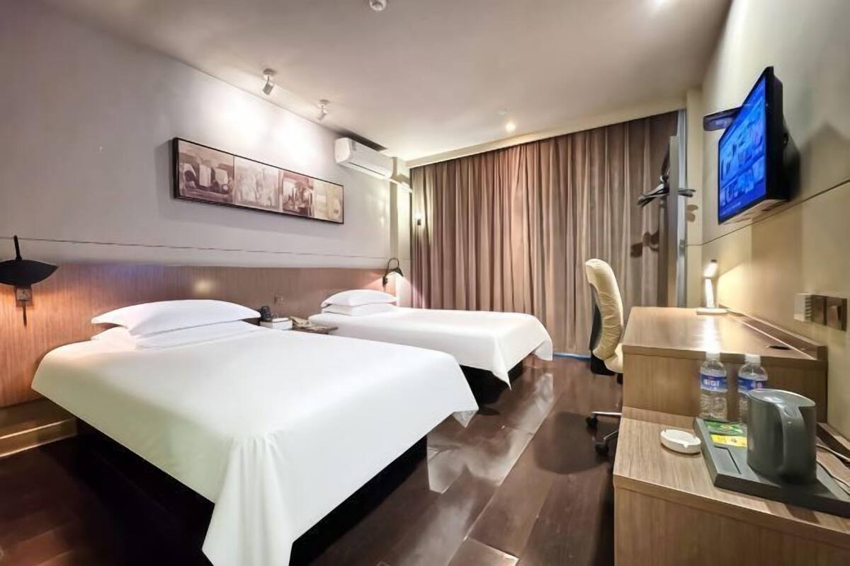 A hotel room with two beds, a desk with a kettle, a TV, and a painting on the wall.