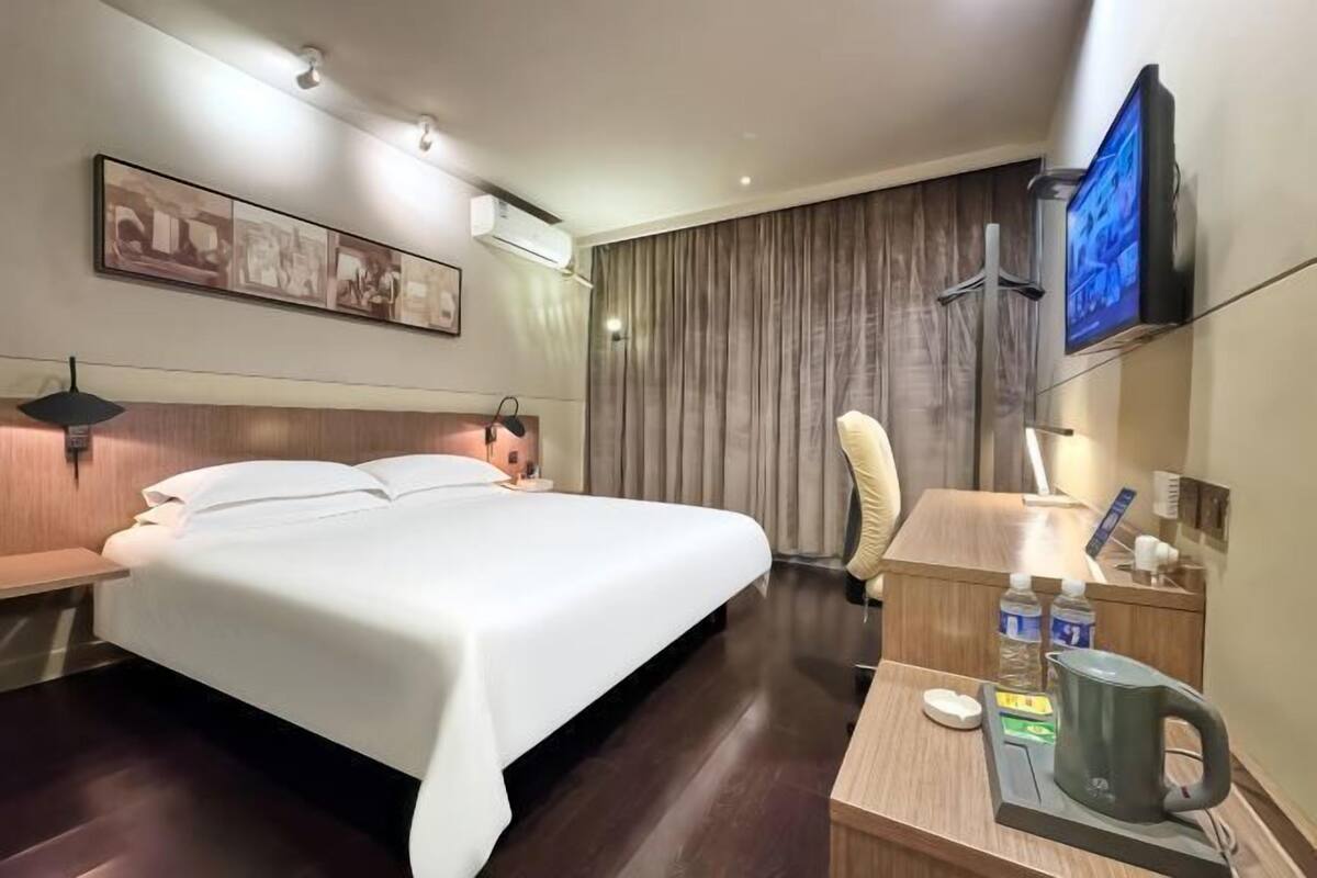 A hotel room with a bed, a desk with a kettle, a television, and a wall-mounted air conditioner.