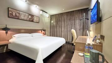 A hotel room with a bed, a desk with a kettle, a television, and a wall-mounted air conditioner.