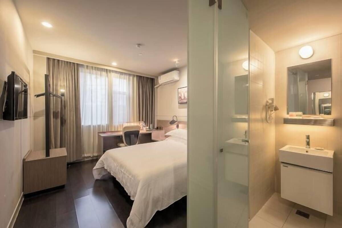 A hotel room with a bed, desk, chair, TV, and a bathroom with a mirror and sink.