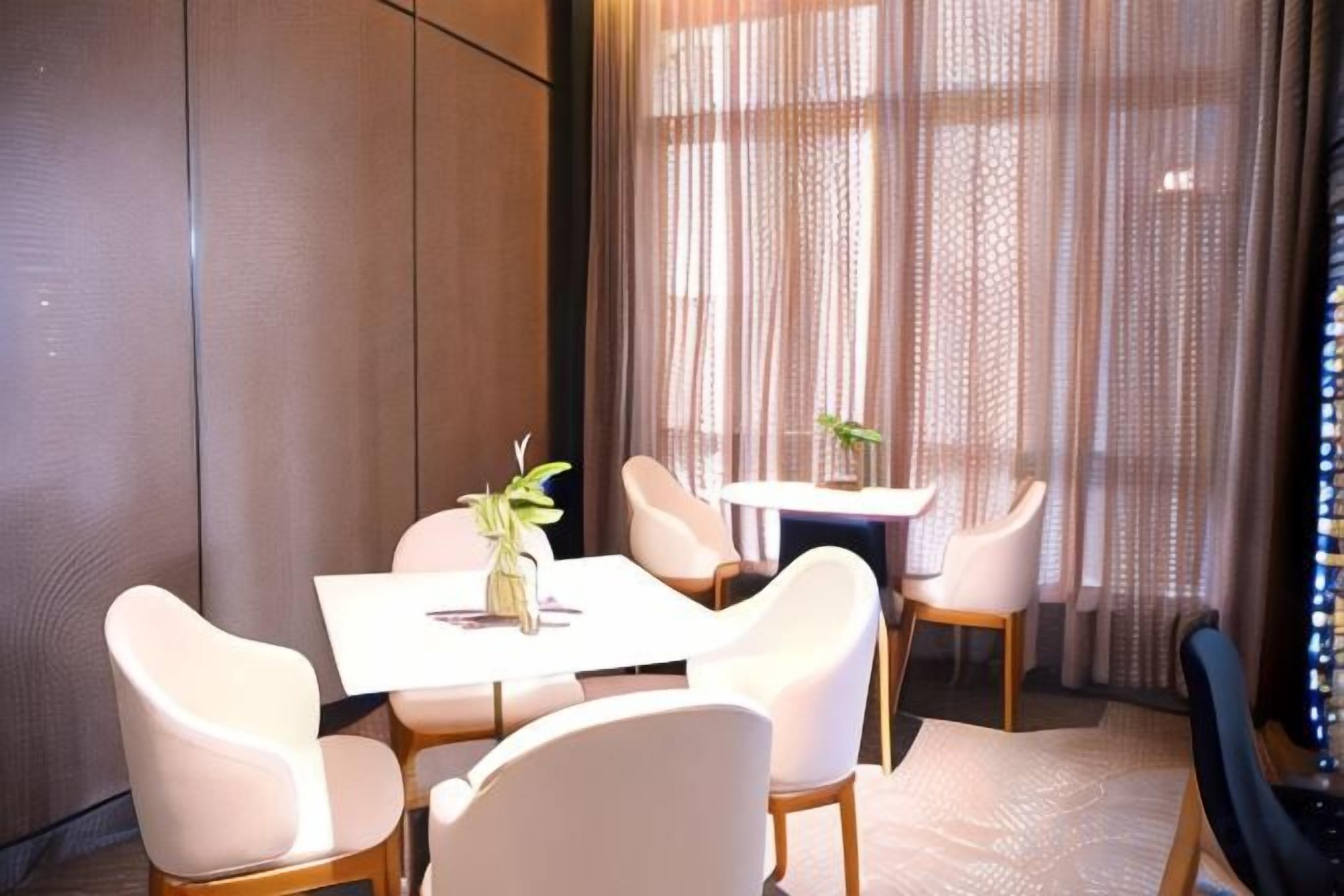A dining area with a table and chairs, a vase with a plant, and a view through sheer curtains.