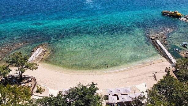Una playa con aguas turquesas cristalinas, una orilla de arena y un muelle con barcos.
