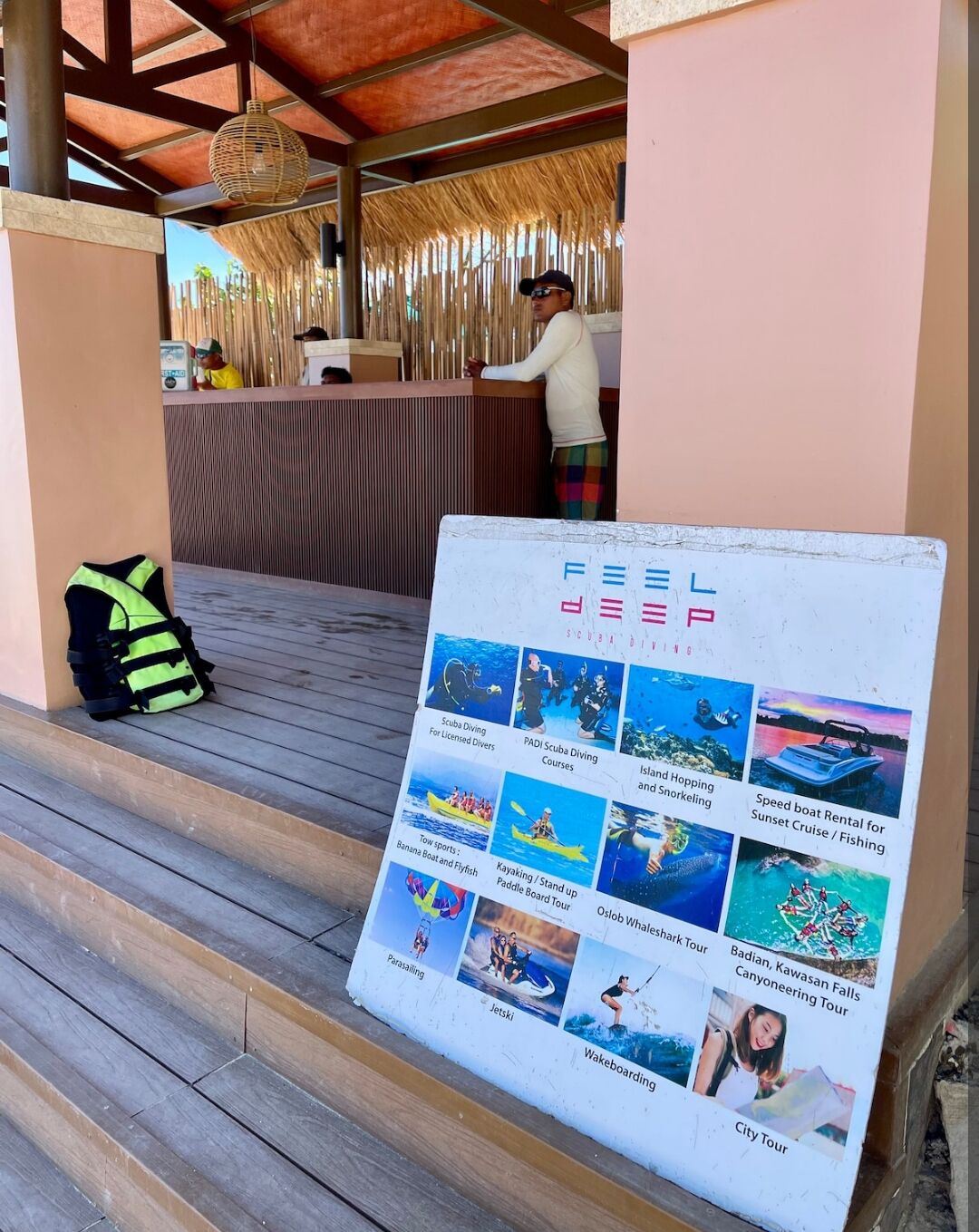 A wooden staircase leading to a covered area with a reception desk. A signboard with various activity options is placed on the steps.