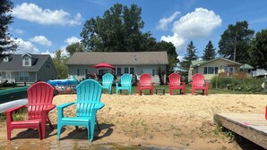 Eine Strandlandschaft mit mehreren Adirondack-Stühlen, einem Holzsteg und einem Haus im Hintergrund.