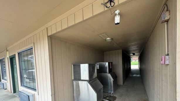 A covered outdoor area with a concrete floor, a metal door, and a security camera mounted on the ceiling.
