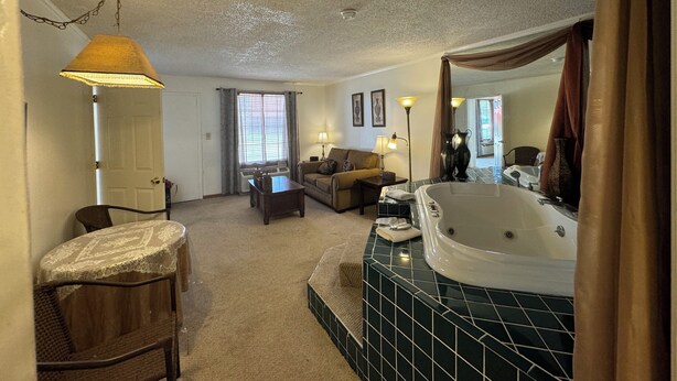 A living room with a sofa, a coffee table, a lamp, and a large window with curtains. - Country Inn (Leesville)