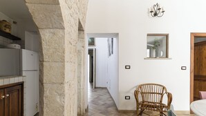 A kitchen with white cabinets, a stone pillar, a wicker chair, and a dining area with a table and chairs. - Appartamento Vintage con Balcone by Wonderful Italy (Ostuni)