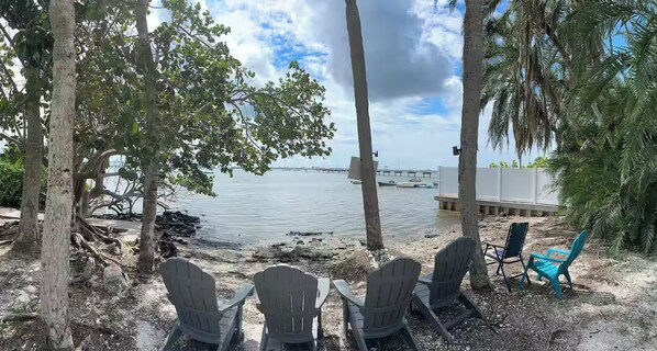A beachside view with chairs and a table, overlooking calm water and a distant bridge. - Waterfront Gulfport Studio w/Shared Private Beach + Walk to Beach & Dining (Gulfport)
