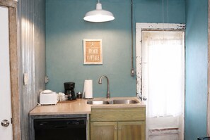 Uma cozinha compacta com pia, cafeteira e torradeira. Há um aviso na parede com a inscrição 'BEACH COTTAGE'.