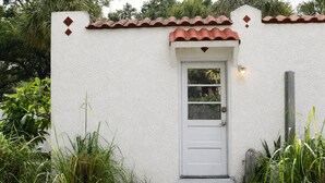 A white exterior wall with a red-tiled roof, a white door with a window, and a small porch area with a blue mat. - Waterfront Gulfport Studio w/Shared Private Beach + Walk to Beach & Dining (Gulfport)