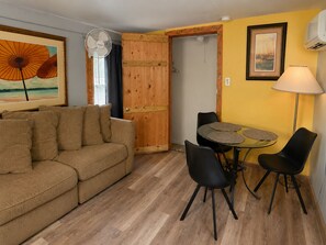Ett mysigt vardagsrum med en soffa, ett matbord med stolar, en fläkt och en dörr som leder till ett annat rum.