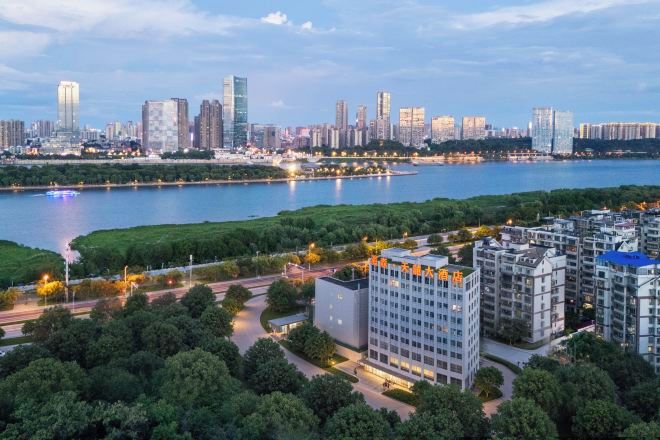 A modern high-rise building with a flat roof and a rectangular shape, surrounded by greenery and a river.