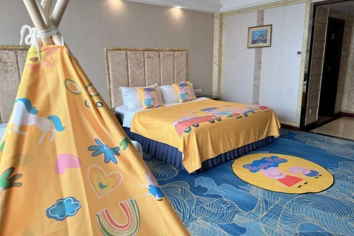 Hotellihuone, jossa on sänky, tyynyjä, värikäs päiväpeite, sininen kuvioitu matto ja telttaa muistuttava rakenne, jossa on sarjakuvakuvioita.