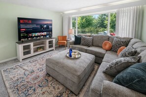 Un salon avec un canapé gris, une table basse, un téléviseur posé sur un meuble et une fenêtre avec des rideaux.