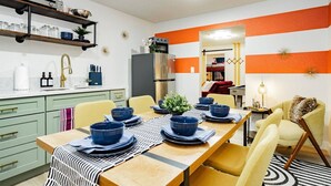 A dining area with a wooden table set for four, surrounded by yellow chairs. There is a kitchen area with a refrigerator and a sink to the left. - Arts District Group Suite | Sleeps 10 + Pool Table (Las Vegas)
