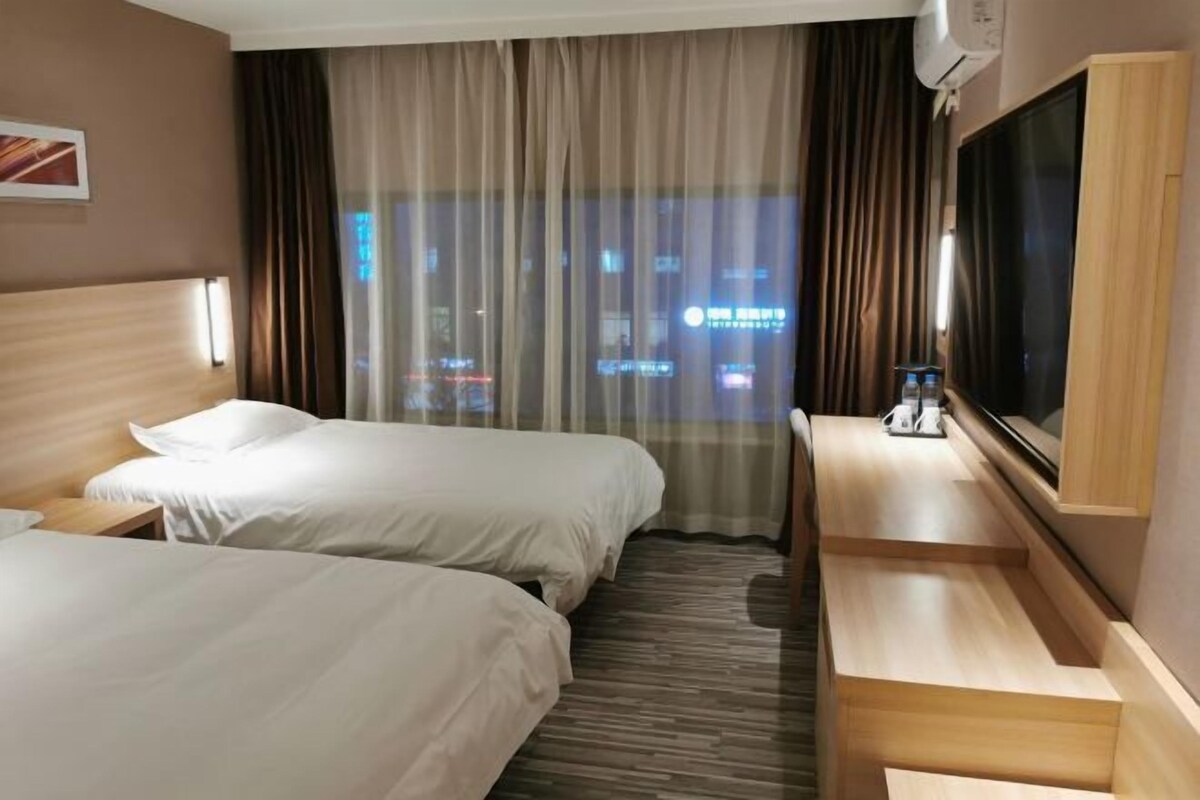 A hotel room with two beds, a desk, a flat-screen TV, and a view of the city at night.