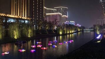 A nighttime view of a river with illuminated boats, a well-lit building with vertical lines, and a bridge in the background.