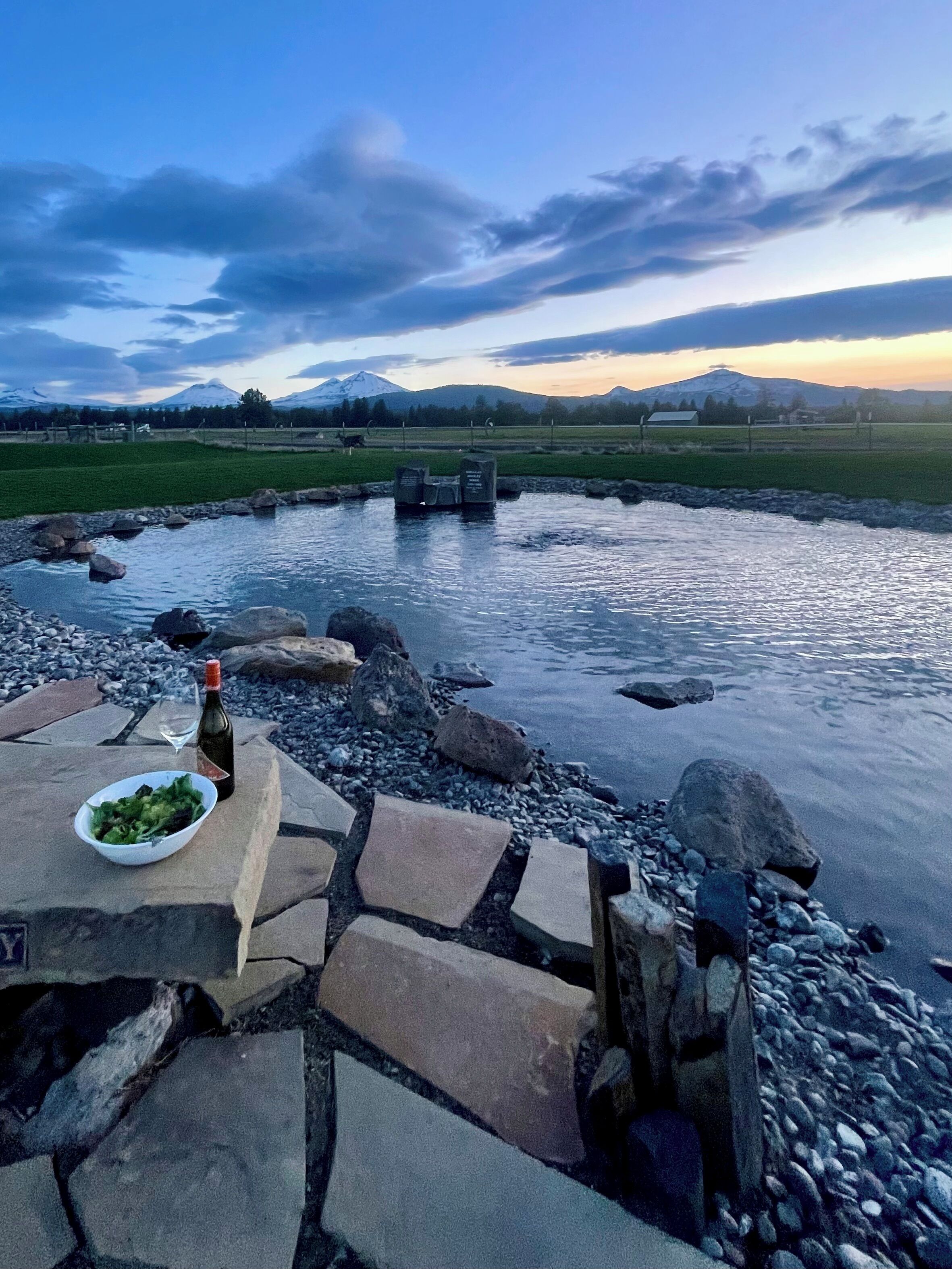 A stone patio with a bowl of salad and a bottle of wine, overlooking a calm river with rocks and a mountainous landscape in the background.
