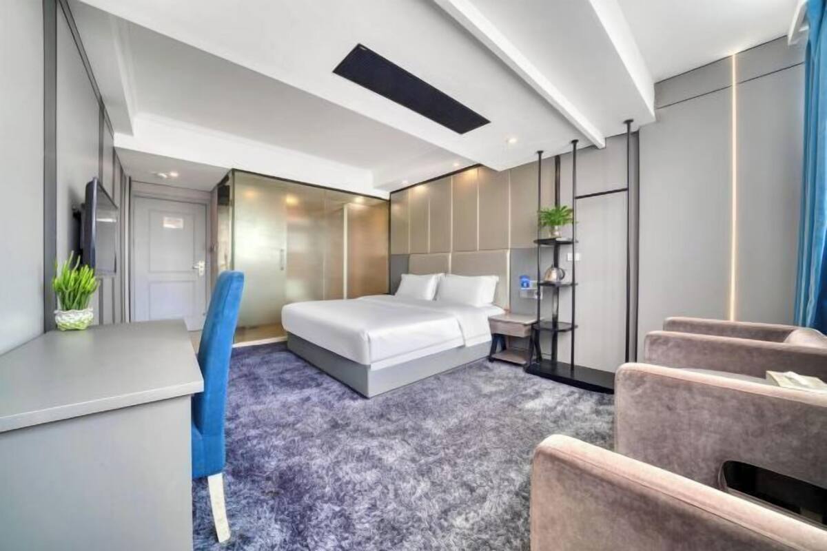 A modern hotel room with a bed, a desk, a chair, and a small seating area.