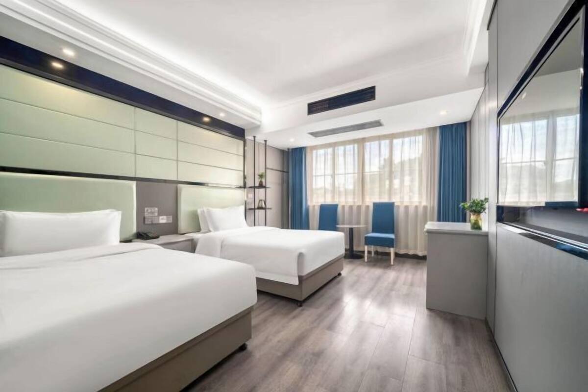 A hotel room with two beds, a small table, chairs, a window with curtains, and a view of the city.