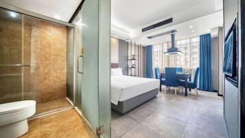 A hotel room with a glass shower enclosure, a bed, a dining table with chairs, and a television.