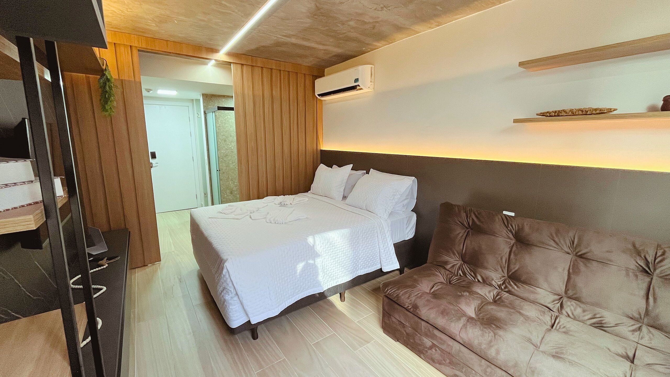Ett hotellrum med en säng, en brun stoppad soffa, en väggpanel av trä och en väggmonterad luftkonditioneringsanläggning.