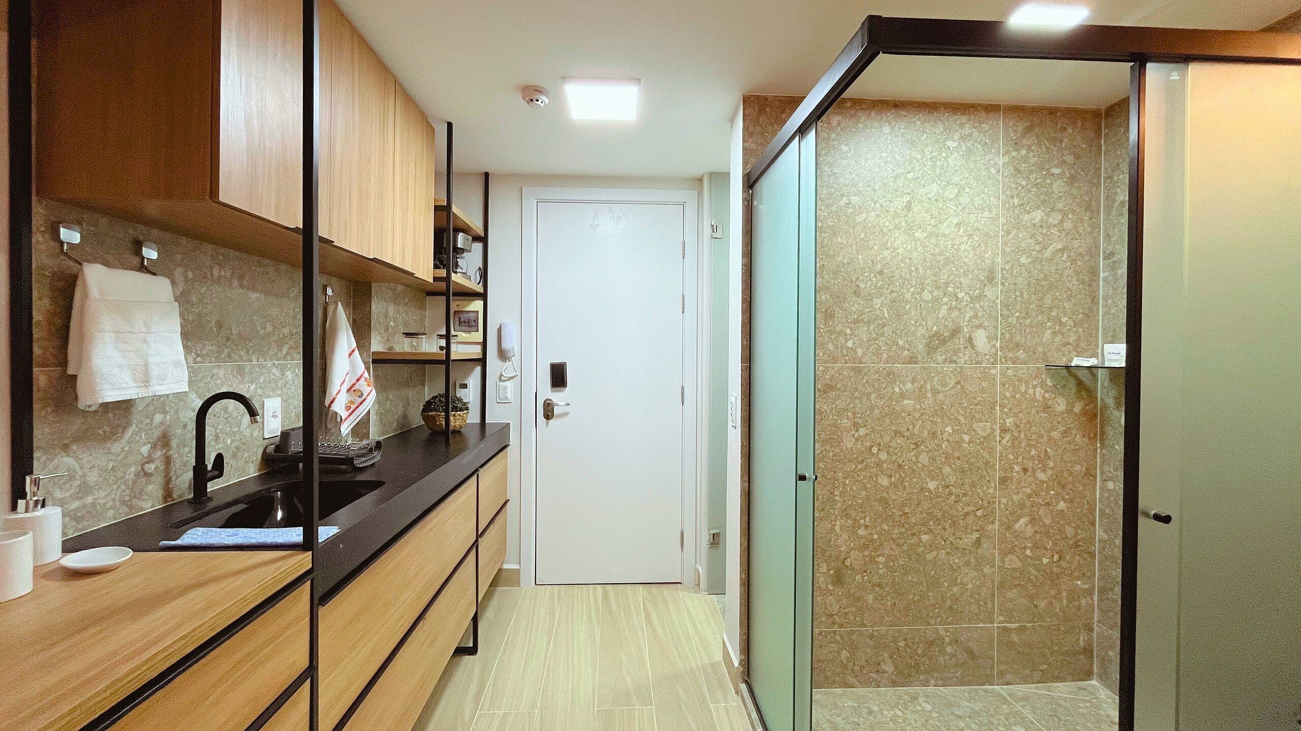 A modern bathroom with a shower, a sink, and a toilet. There is a towel hanging on the wall and a small shelf with toiletries.