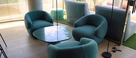 Un espace de détente moderne avec des fauteuils turquoise et une table en verre, de grandes fenêtres et une vue sur l’extérieur.