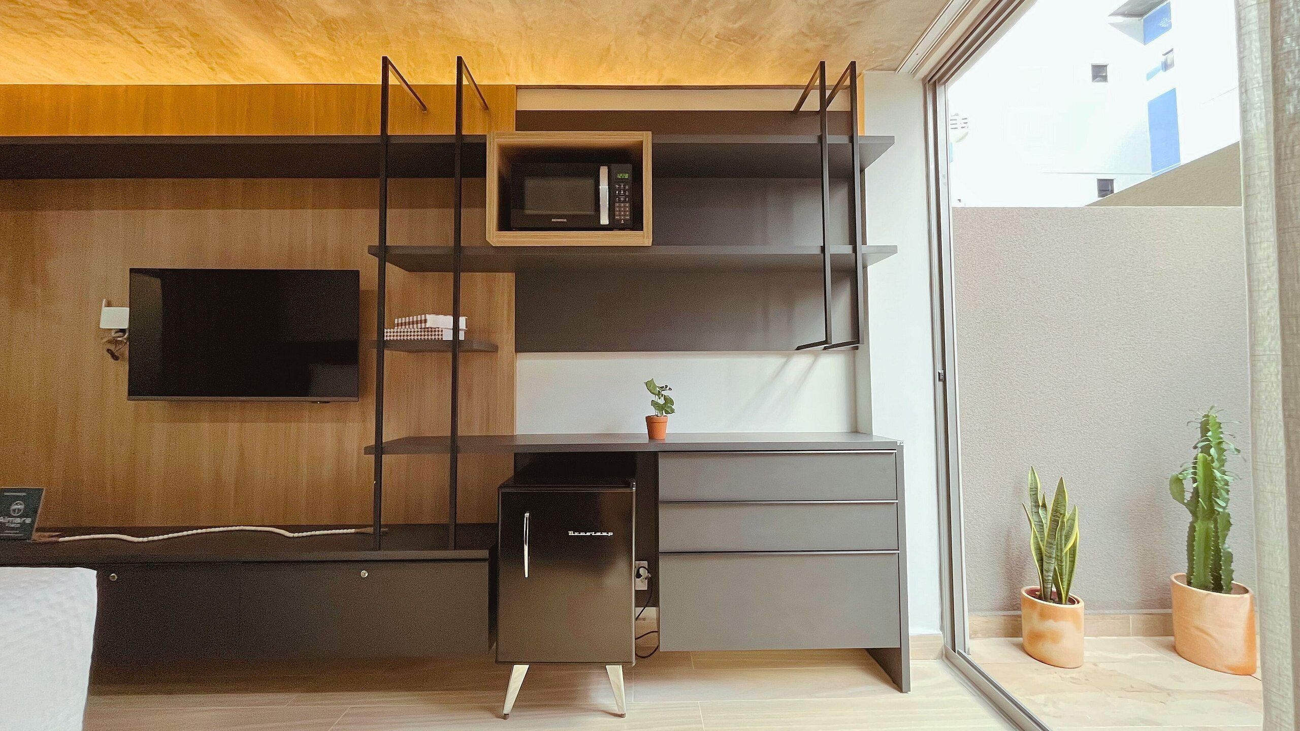 A compact kitchen with a microwave, a small refrigerator, and a TV mounted on the wall.