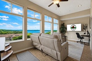 A room with a massage chair, a desk, a computer, and a large window with a view of the ocean and greenery.