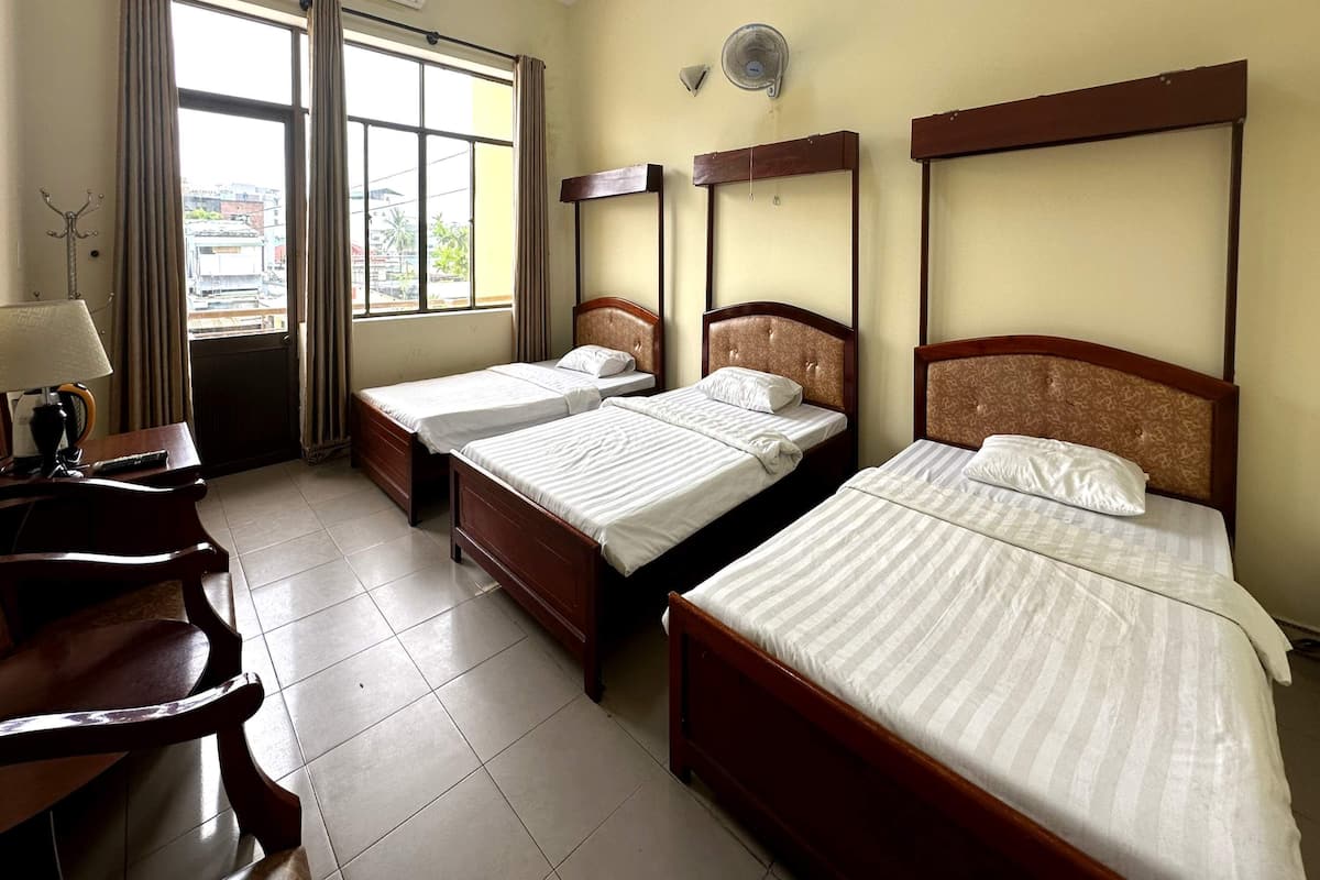 A hotel room with three single beds, a desk with a lamp, a chair, and a window with curtains.