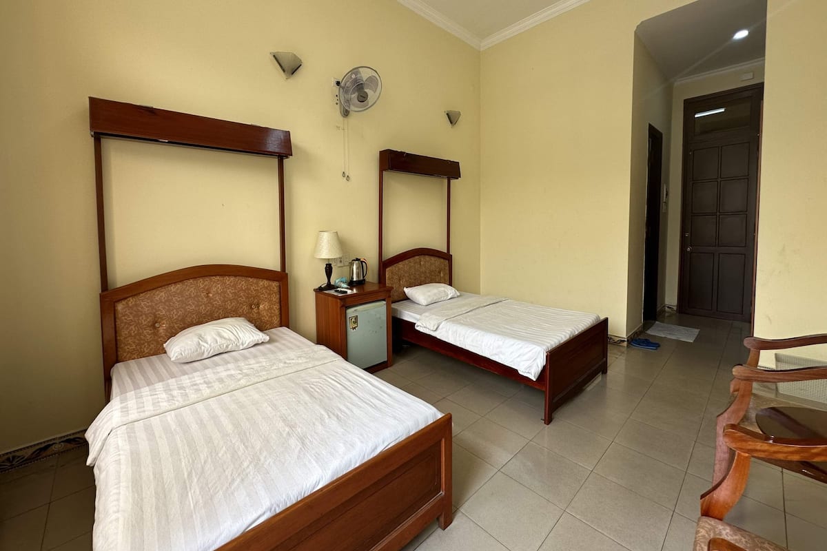 A hotel room with two wooden beds, a small bedside table with a lamp, a wall-mounted fan, and a door leading to another room.