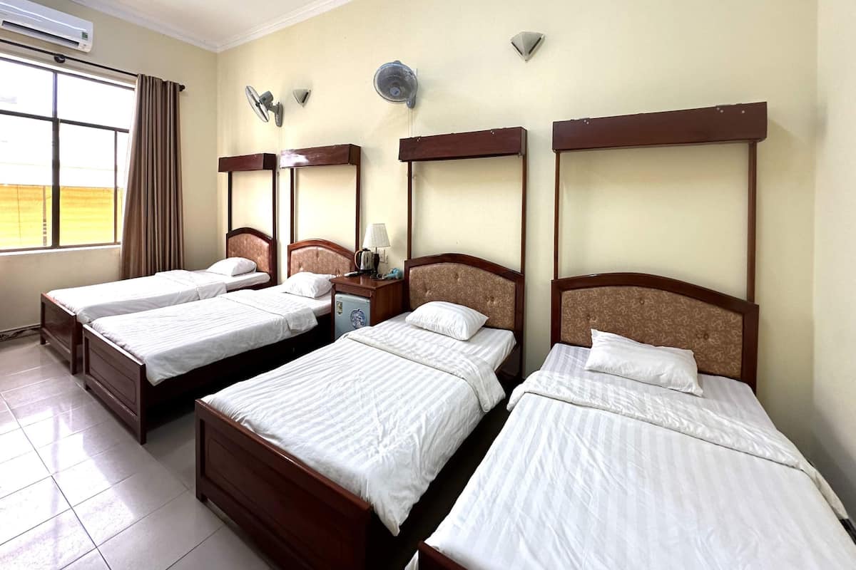 A hotel room with four single beds, a small table with a lamp, an air conditioner, and a window with curtains.