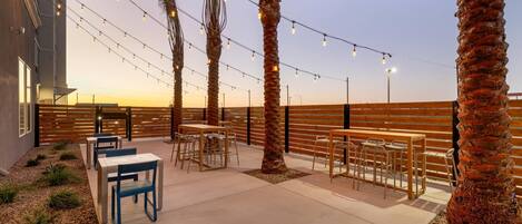 An outdoor patio with tables and chairs, palm trees, string lights, and a wooden fence.